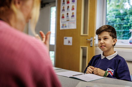 Boy sat with a teacher doing one to one work. You can see an open door