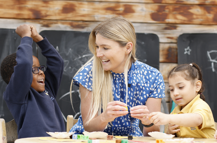 A young girl and boy sat outside with a teacher infront of blackboards